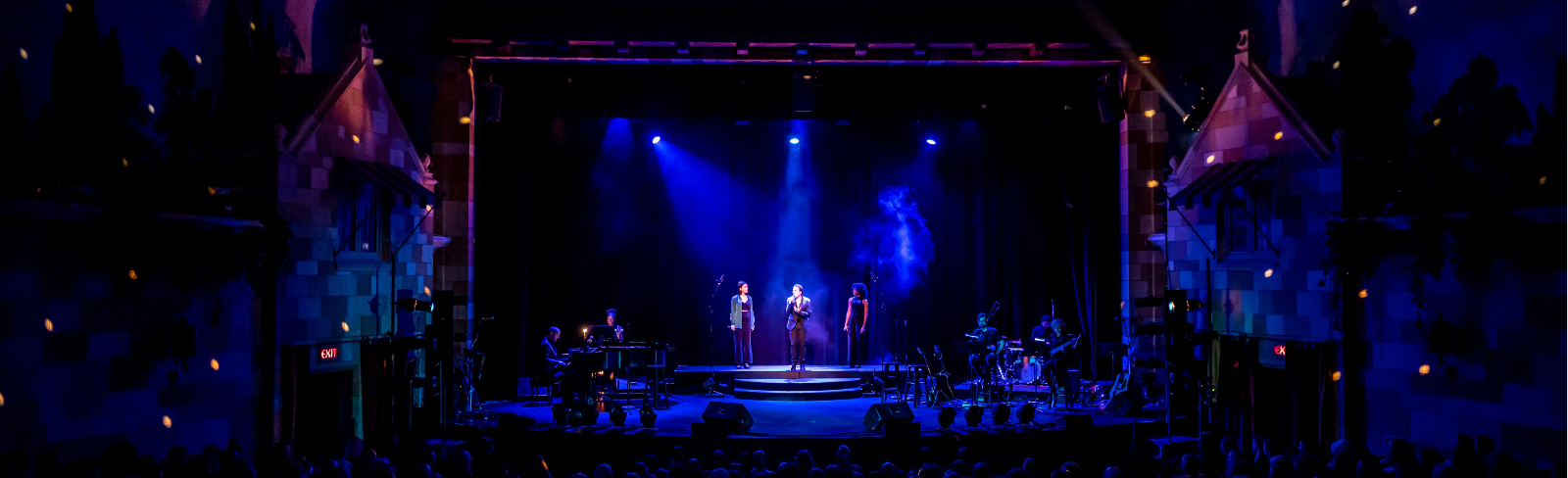Image of three performers on stage singing into microphones singing to a full audience